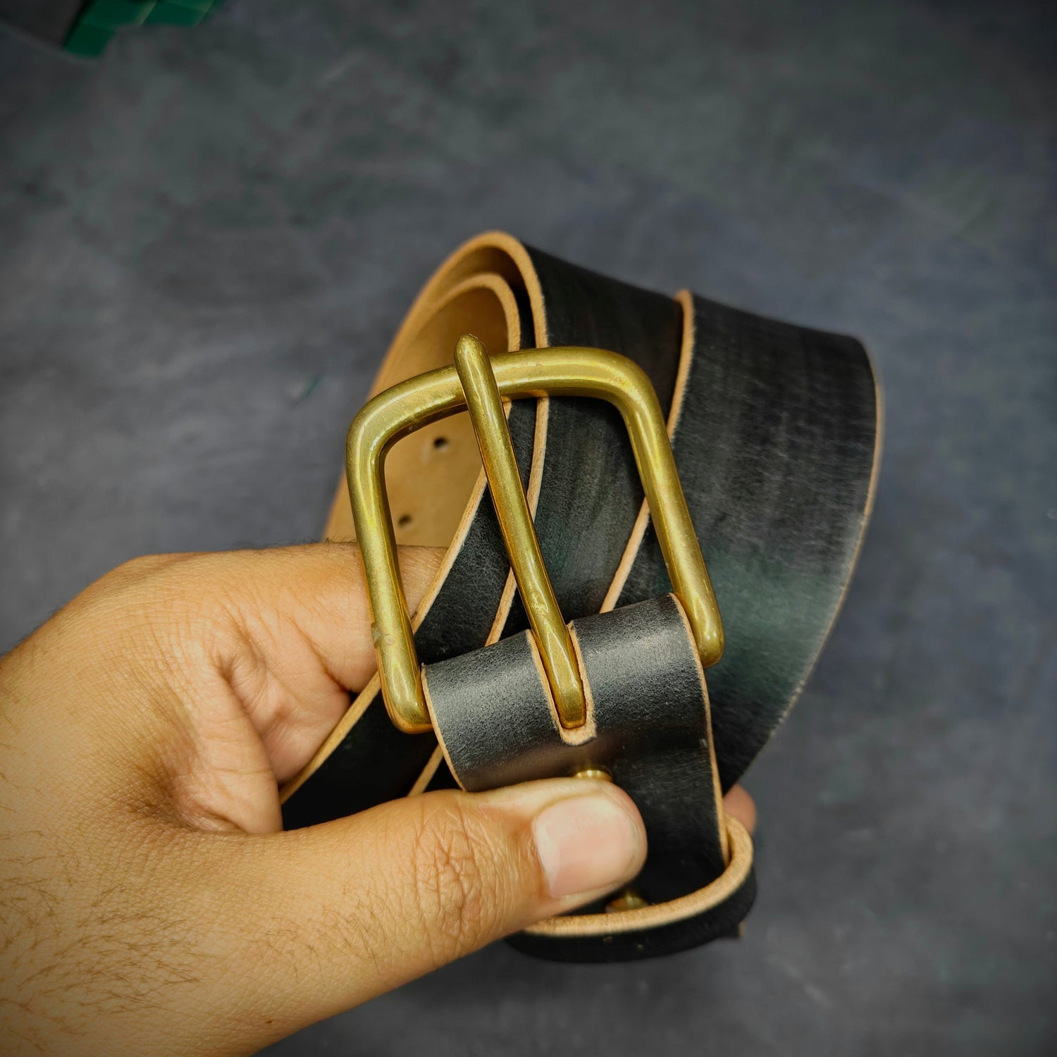 A close-up of a hand holding a black leather belt with a 35mm, brass buckle against a dark textured background.