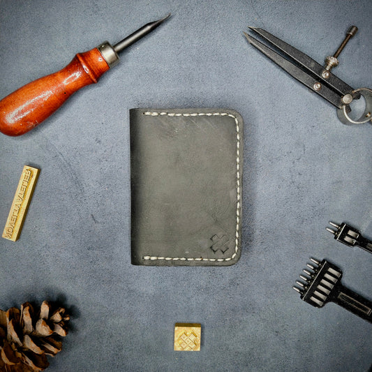 The Copperfield full grain leather bifold wallet by Novelty Atelier closed view highlighting slim and rugged design. Handcrafted from Black and Brown ethically sourced leather, saddle stitched in off white thread. Set against a textured background with branding tools and pine cone, perfect for minimalist EDC style.

