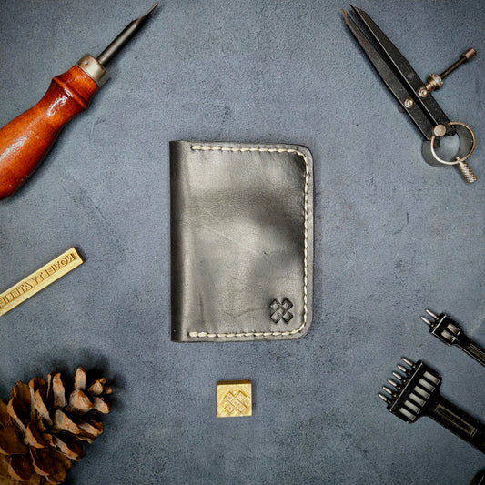 The Copperfield full grain leather bifold wallet by Novelty Atelier closed view highlighting slim and rugged design. Handcrafted from Black and Green ethically sourced leather, saddle stitched in off white thread. Set against a textured background with branding tools and pine cone, perfect for minimalist EDC style.
