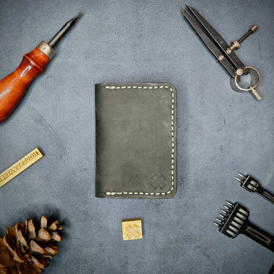 The Copperfield full grain leather bifold wallet by Novelty Atelier closed view highlighting slim and rugged design. Handcrafted from Black and Purple ethically sourced leather, saddle stitched in off white thread. Set against a textured background with branding tools and pine cone, perfect for minimalist EDC style.
