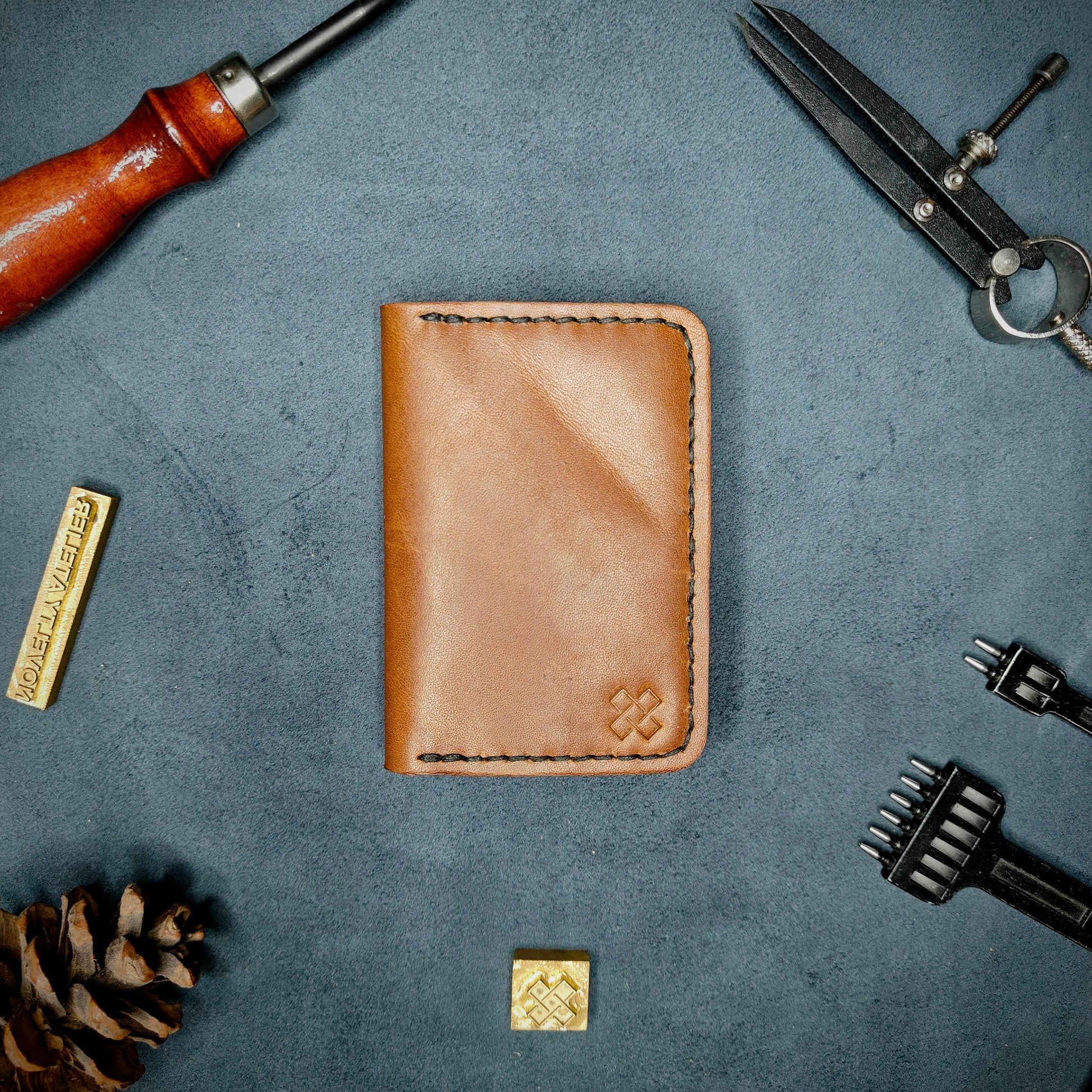 The Copperfield full grain leather bifold wallet by Novelty Atelier closed view highlighting slim and rugged design. Handcrafted from Brown ethically sourced leather, saddle stitched in black thread. Set against a textured background with branding tools and pine cone, perfect for minimalist EDC style.
