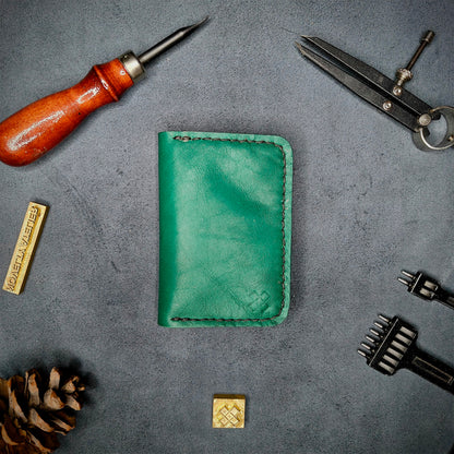 The Copperfield full grain leather bifold wallet by Novelty Atelier closed view highlighting slim and rugged design. Handcrafted from Green ethically sourced leather, saddle stitched in black thread. Set against a textured background with branding tools and pine cone, perfect for minimalist EDC style.
