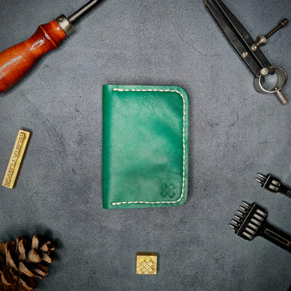 The Copperfield full grain leather bifold wallet by Novelty Atelier closed view highlighting slim and rugged design. Handcrafted from Green ethically sourced leather, saddle stitched in off white thread. Set against a textured background with branding tools and pine cone, perfect for minimalist EDC style.
