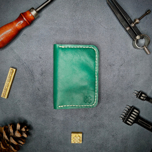 The Copperfield full grain leather bifold wallet by Novelty Atelier closed view highlighting slim and rugged design. Handcrafted from Green ethically sourced leather, saddle stitched in off white thread. Set against a textured background with branding tools and pine cone, perfect for minimalist EDC style.
