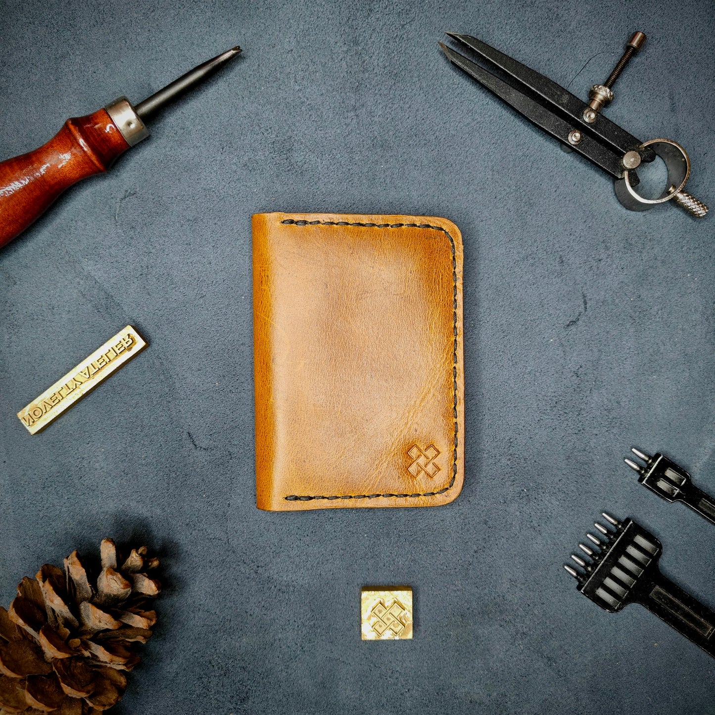 The Copperfield full grain leather bifold wallet by Novelty Atelier closed view highlighting slim and rugged design. Handcrafted from Mustard yellow ethically sourced leather, saddle stitched in black thread. Set against a textured background with branding tools and pine cone, perfect for minimalist EDC style.
