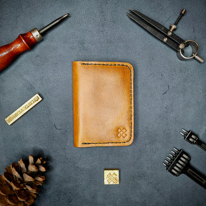 The Copperfield full grain leather bifold wallet by Novelty Atelier closed view highlighting slim and rugged design. Handcrafted from Mustard yellow ethically sourced leather, saddle stitched in black thread. Set against a textured background with branding tools and pine cone, perfect for minimalist EDC style.
