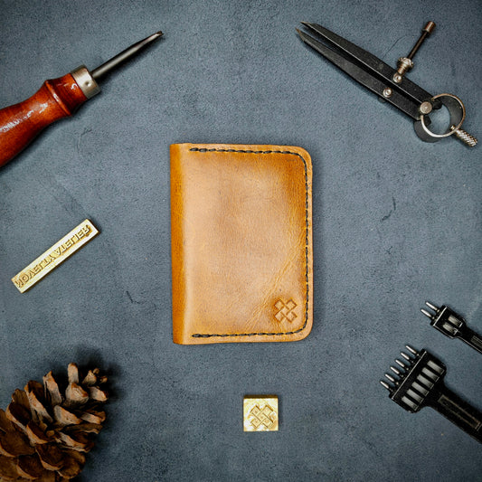 The Copperfield full grain leather bifold wallet by Novelty Atelier closed view highlighting slim and rugged design. Handcrafted from Mustard yellow ethically sourced leather, saddle stitched in black thread. Set against a textured background with branding tools and pine cone, perfect for minimalist EDC style.
