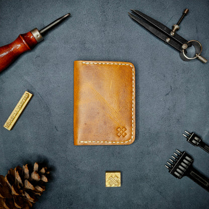 The Copperfield full grain leather bifold wallet by Novelty Atelier closed view highlighting slim and rugged design. Handcrafted from Mustard yellow ethically sourced leather, saddle stitched in off white  thread. Set against a textured background with branding tools and pine cone, perfect for minimalist EDC style.
