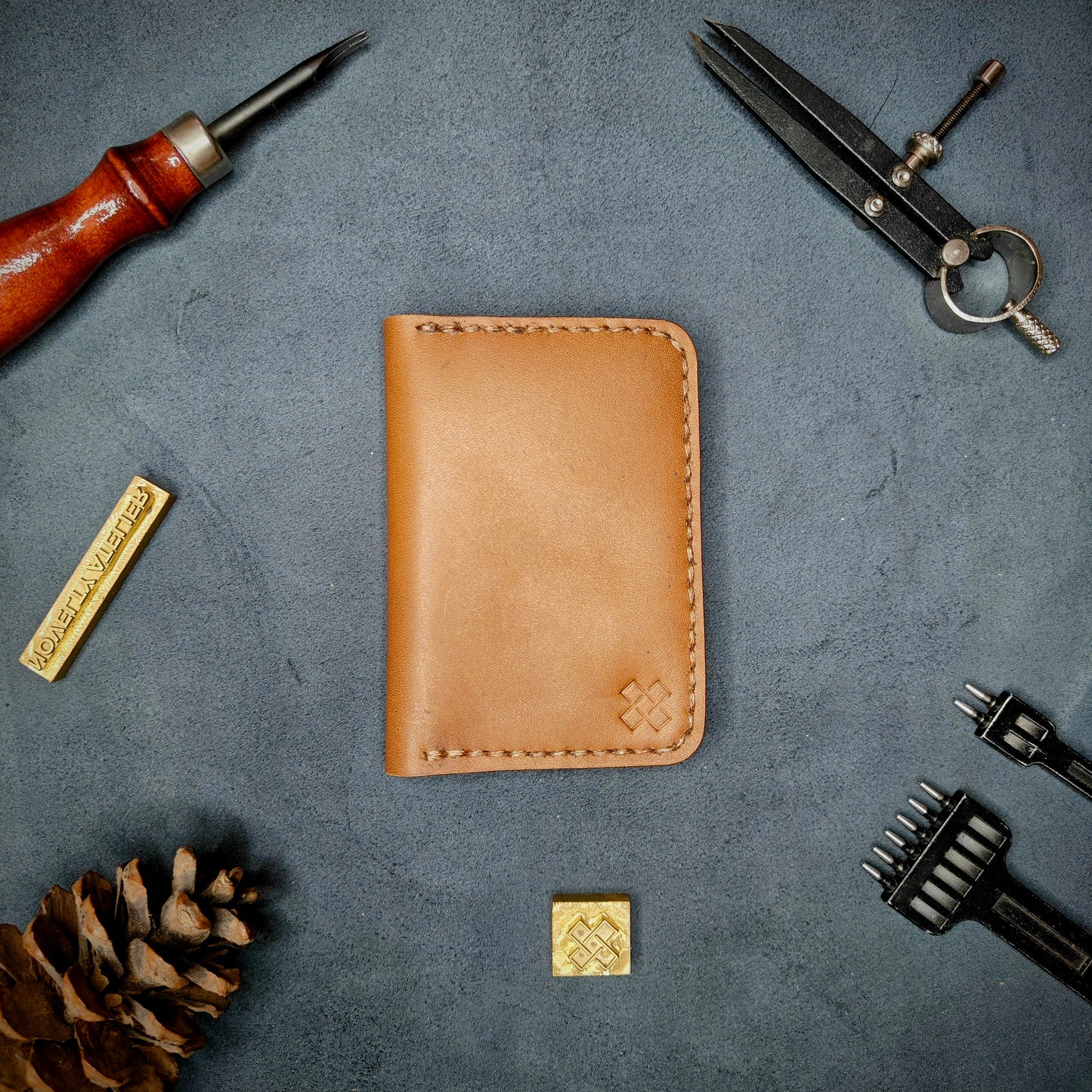 The Copperfield full grain leather bifold wallet by Novelty Atelier closed view highlighting slim and rugged design. Handcrafted from Tann ethically sourced leather, saddle stitched in Brown thread. Set against a textured background with branding tools and pine cone, perfect for minimalist EDC style.
