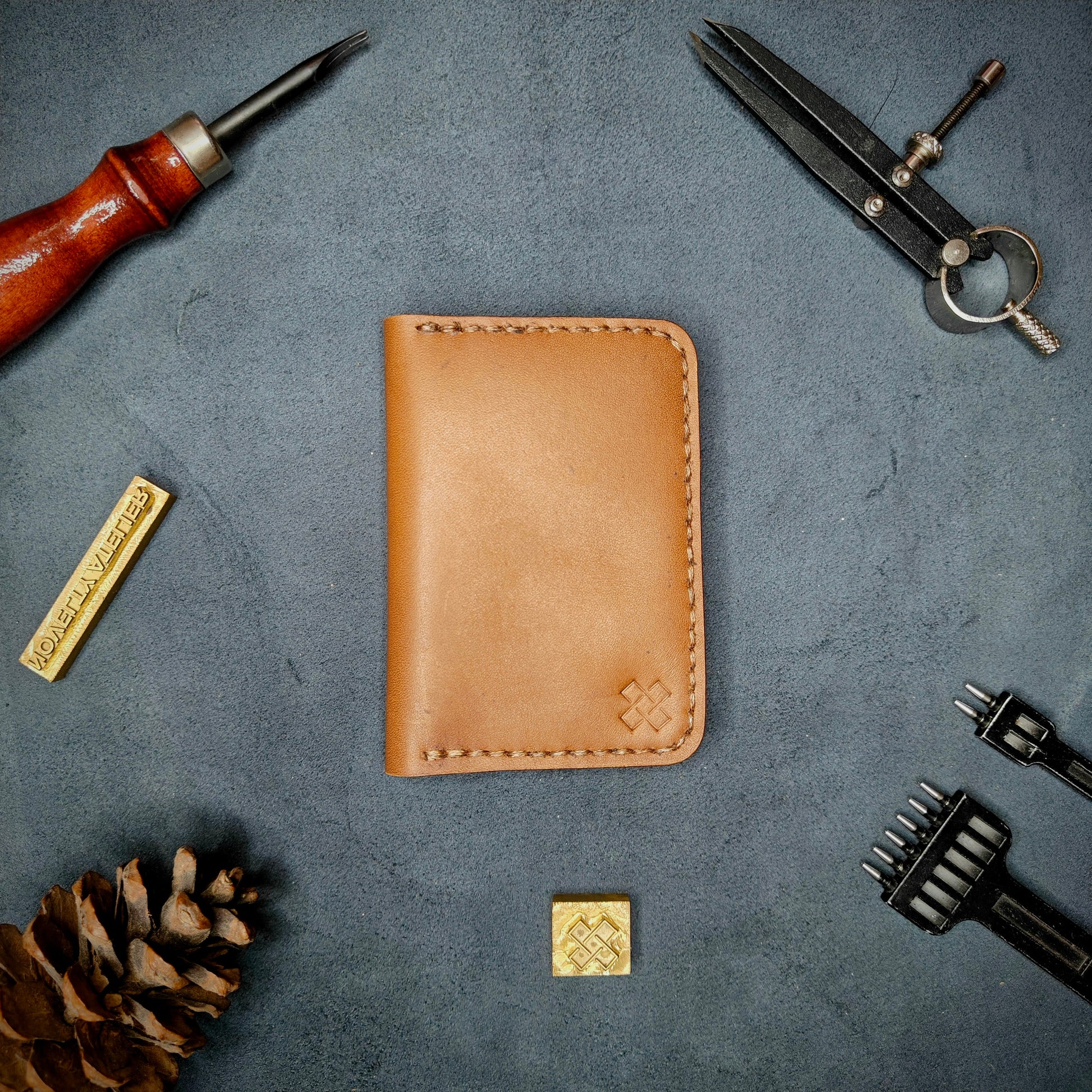 The Copperfield full grain leather bifold wallet by Novelty Atelier closed view highlighting slim and rugged design. Handcrafted from Tann ethically sourced leather, saddle stitched in Brown thread. Set against a textured background with branding tools and pine cone, perfect for minimalist EDC style.
