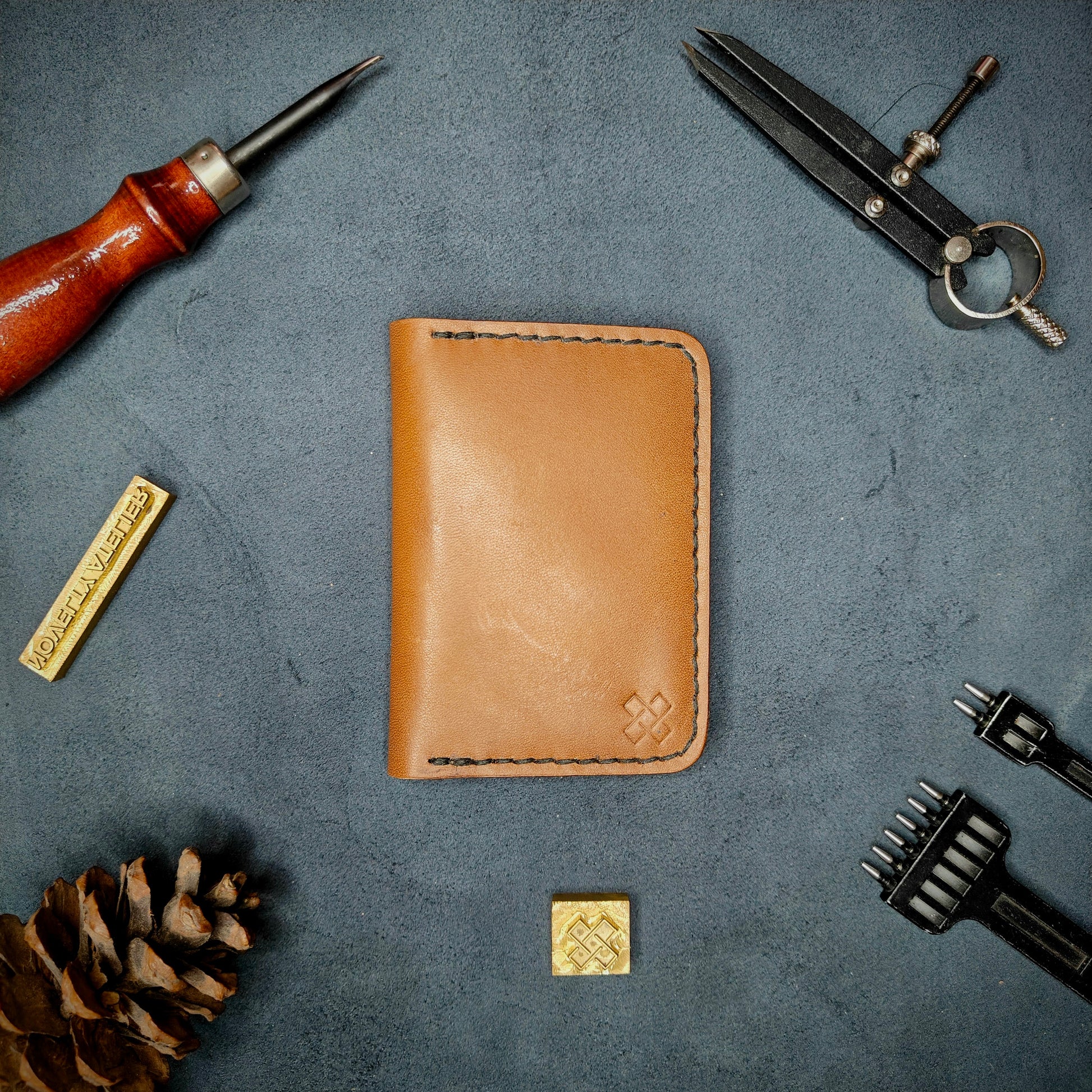 The Copperfield full grain leather bifold wallet by Novelty Atelier closed view highlighting slim and rugged design. Handcrafted from Tann ethically sourced leather, saddle stitched in black thread. Set against a textured background with branding tools and pine cone, perfect for minimalist EDC style.
