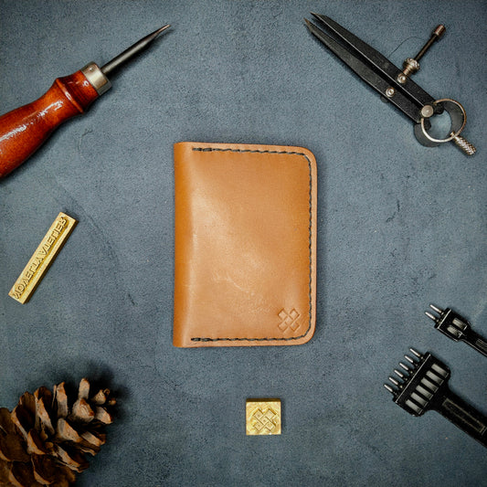 The Copperfield full grain leather bifold wallet by Novelty Atelier closed view highlighting slim and rugged design. Handcrafted from Tann ethically sourced leather, saddle stitched in black thread. Set against a textured background with branding tools and pine cone, perfect for minimalist EDC style.
