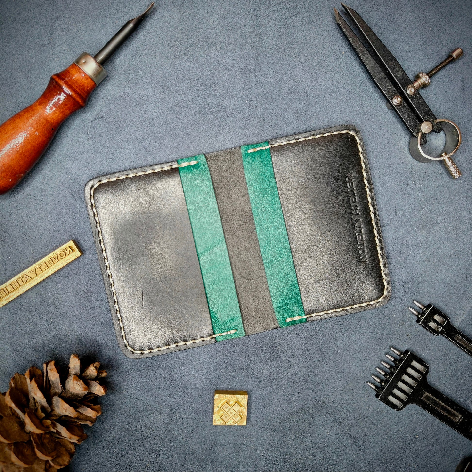 The Copperfield handmade leather card holder by Novelty Atelier open flat view revealing 4 card slots, crafted from ethically sourced Black and Green full grain leather with off white saddle stitching. Displayed on suede with brass stamp, leatherworking tools, and pine cone for EDC inspired composition. A durable, minimalist wallet for everyday carry.
