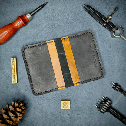 The Copperfield handmade leather card holder by Novelty Atelier open flat view revealing 4 card slots, crafted from ethically sourced Black and Mustard full grain leather with black thread saddle stitching. Displayed on suede with brass stamp, leatherworking tools, and pine cone for EDC inspired composition. A durable, minimalist wallet for everyday carry.
