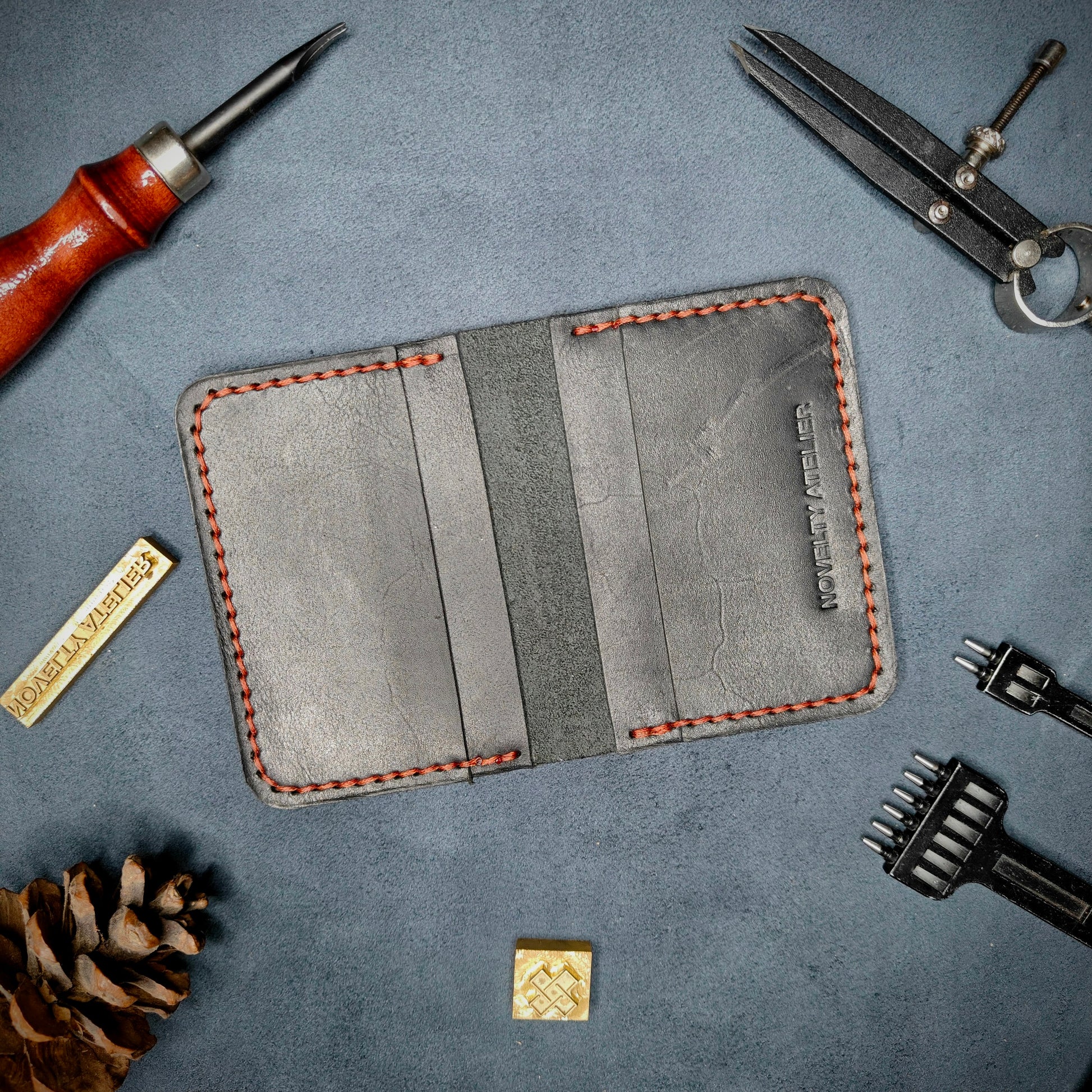 The Copperfield handmade leather card holder by Novelty Atelier open flat view revealing 4 card slots, crafted from ethically sourced Black full grain leather with Red thread saddle stitching. Displayed on suede with brass stamp, leather working tools, and pine cone for EDC inspired composition. A durable, minimalist wallet for everyday carry.
