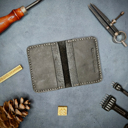 The Copperfield handmade leather card holder by Novelty Atelier open flat view revealing 4 card slots, crafted from ethically sourced Black full grain leather with off white  saddle stitching. Displayed on suede with brass stamp, leatherworking tools, and pine cone for EDC inspired composition. A durable, minimalist wallet for everyday carry.
