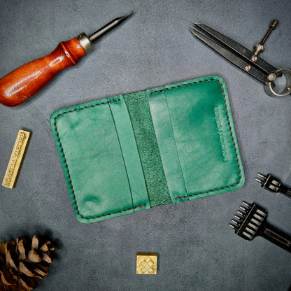 The Copperfield handmade leather card holder by Novelty Atelier open flat view revealing 4 card slots, crafted from ethically sourced Green full grain leather with black thread saddle stitching. Displayed on suede with brass stamp, leatherworking tools, and pine cone for EDC inspired composition. A durable, minimalist wallet for everyday carry.
