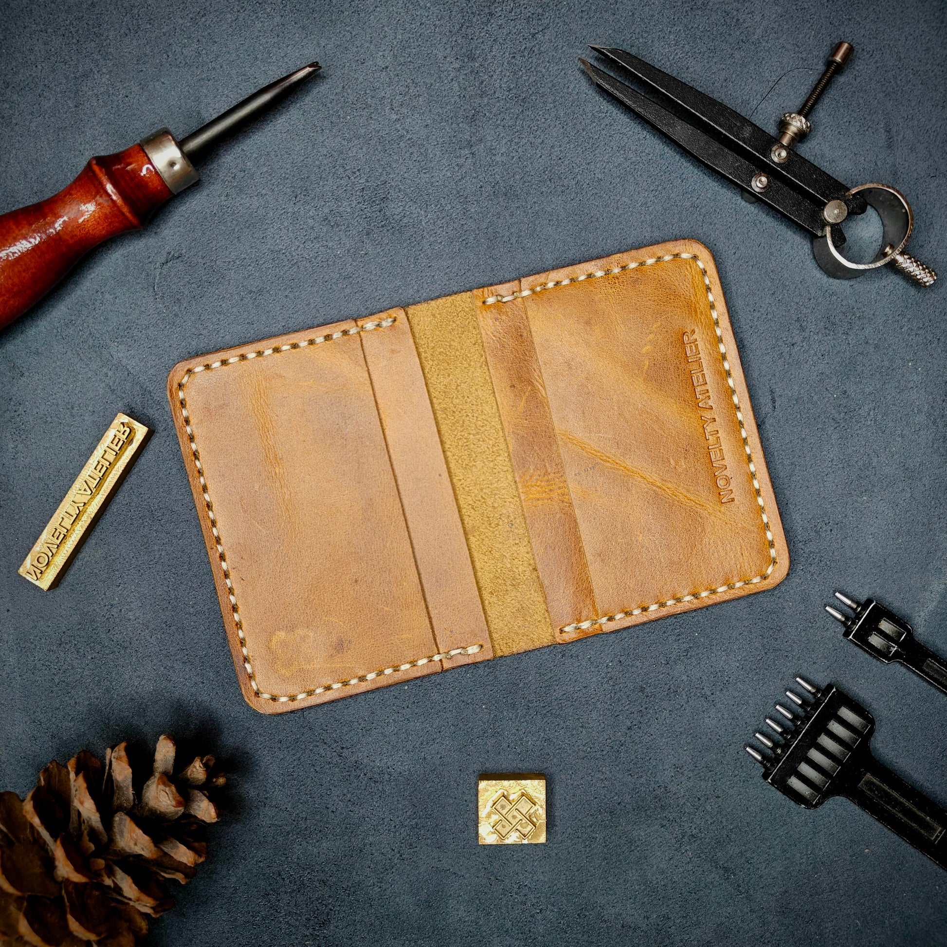 The Copperfield handmade leather card holder by Novelty Atelier open flat view revealing 4 card slots, crafted from ethically sourced Mustard yellow full grain leather with off white  saddle stitching. Displayed on suede with brass stamp, leatherworking tools, and pine cone for EDC inspired composition. A durable, minimalist wallet for everyday carry.
