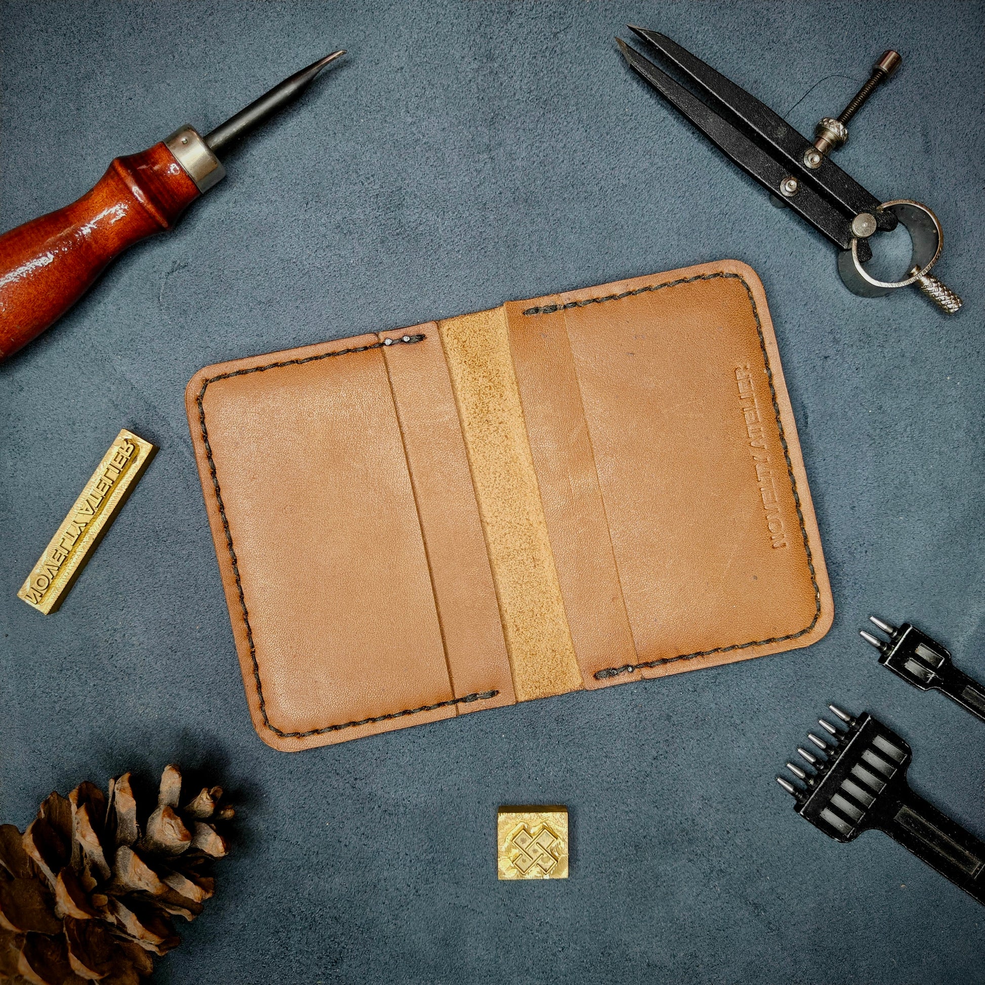 The Copperfield handmade leather card holder by Novelty Atelier open flat view revealing 4 card slots, crafted from ethically sourced Tann full grain leather with black thread saddle stitching. Displayed on suede with brass stamp, leatherworking tools, and pine cone for EDC inspired composition. A durable, minimalist wallet for everyday carry.
