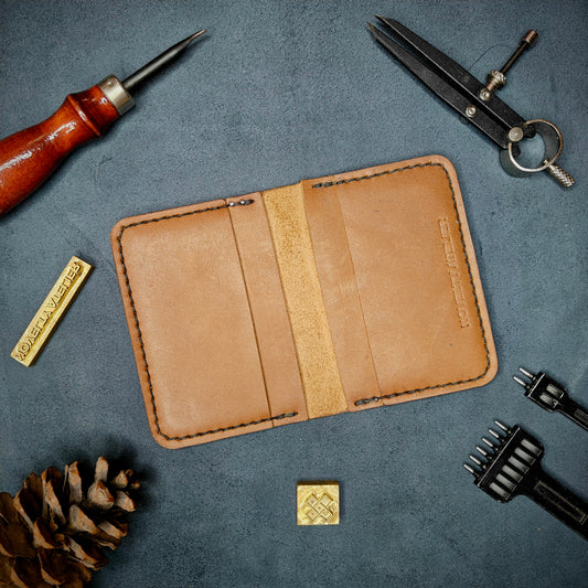 The Copperfield handmade leather card holder by Novelty Atelier open flat view revealing 4 card slots, crafted from ethically sourced Tann full grain leather with black thread saddle stitching. Displayed on suede with brass stamp, leatherworking tools, and pine cone for EDC inspired composition. A durable, minimalist wallet for everyday carry.
