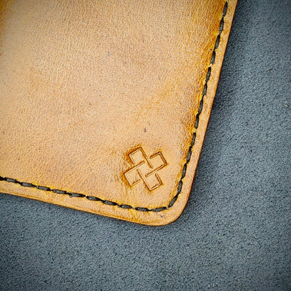 Zoomed in view of Novelty Atelier’s signature insignia embossed on The Copperfield minimalist leather card holder. Hand stitched in black thread using traditional saddle technique. Crafted from Mustard yellow full grain leather, symbolizing timeless durability and ethical sophistication.
